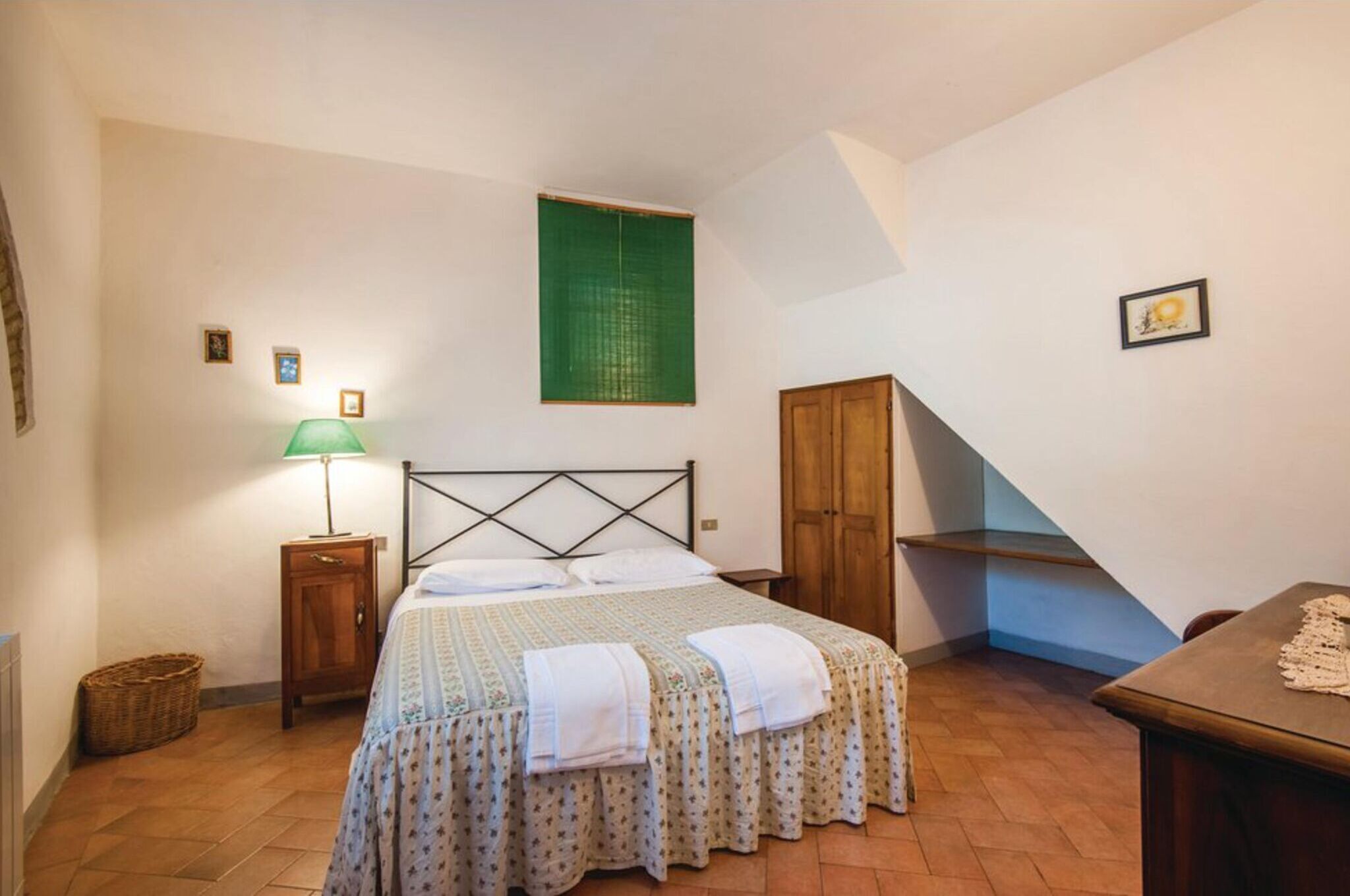 Een slaapkamer met een bed, nachtkastje, lamp, groen raam, houten garderobe en een tafel.