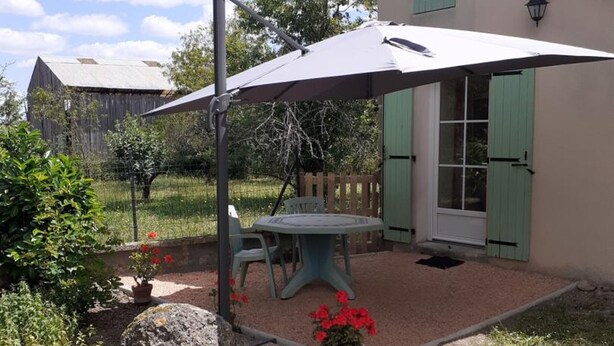 An outdoor patio with a table and chairs under a large umbrella, surrounded by a garden with potted plants and a large rock.