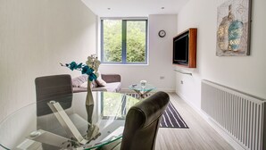 Un salon moderne avec une table à manger en verre, des fauteuils en cuir, un canapé, un téléviseur fixé au mur et une grande fenêtre donnant sur un paysage arboré.