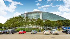 Een modern stadion met een glazen gevel en een gebogen dak, omringd door geparkeerde auto's en bomen.