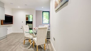 Een moderne eetruimte met een glazen tafel en beklede stoelen, een flatscreen televisie en een groot raam met uitzicht op groen.