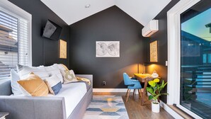 En moderne stue med en grå sofa, en blå lænestol og et lille bord med en lampe. Der er et stort vindue med udsigt til bygninger.