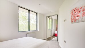 Un dormitorio con un ventanal grande, una cama, un reposapi és rojo y una puerta que da a otra habitación.