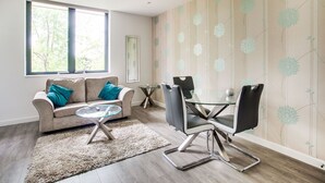 Una sala de estar moderna con un sofá, una mesa de centro de vidrio y un comedor con una mesa y sillas de vidrio. Hay un ventanal que proporciona luz natural y vistas a árboles.