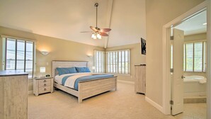 Une chambre à coucher avec un lit, des tables de chevet, un ventilateur de plafond et une salle de bain visible à travers une porte ouverte.