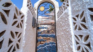 A doorway with intricate white carvings and a stained glass window depicting a sun, clouds, palm tree, and waves.