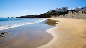 A beach with clear blue water, sandy shore, and white buildings on a hill in the background.