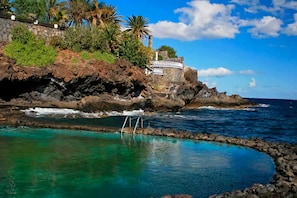 A coastal scene with a clear pool, rocky shoreline, and a building with a white railing. - Belvilla by OYO Holiday home in Arico El Nuevo (Arico El Nuevo)