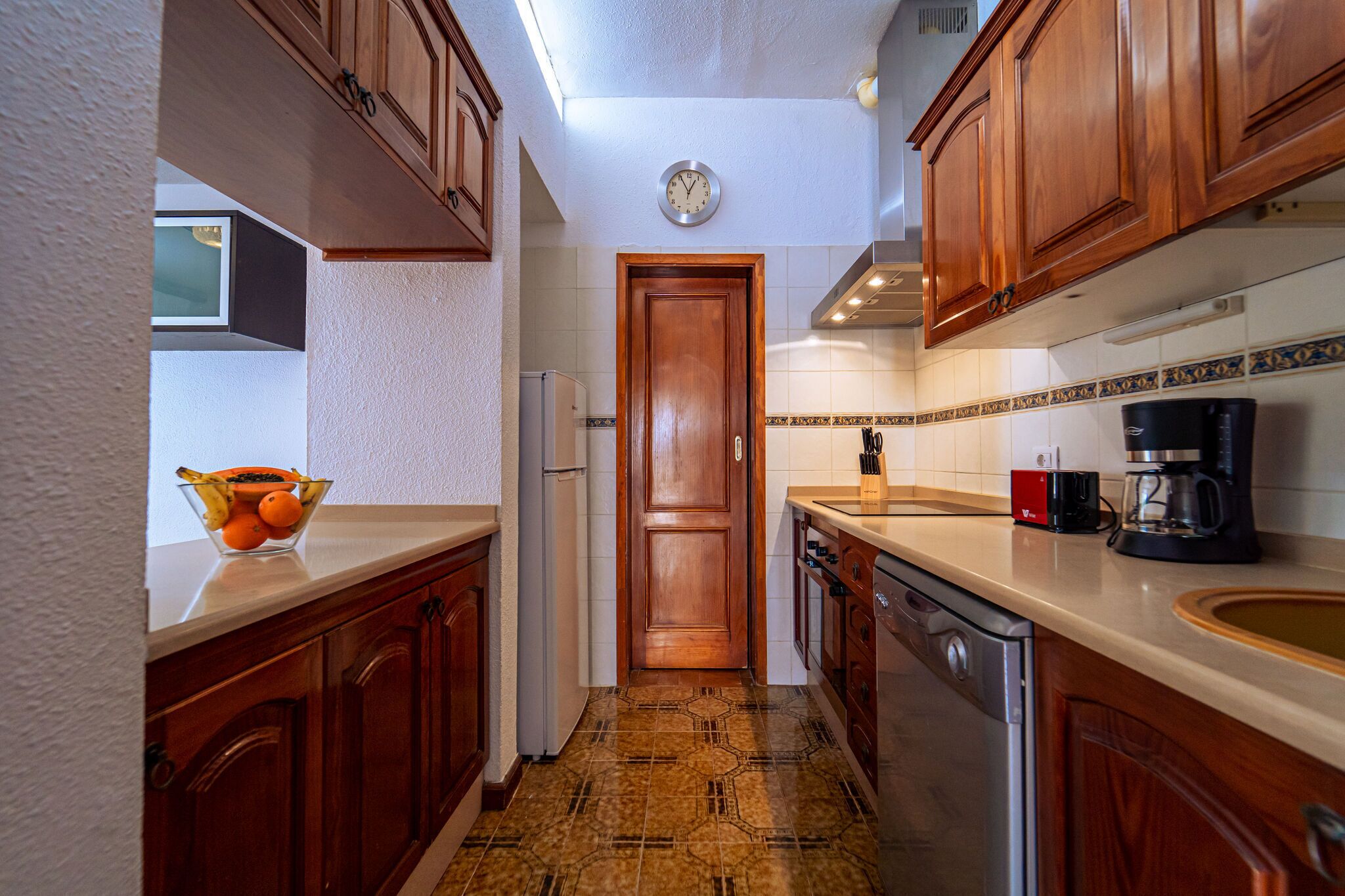 A kitchen with wooden cabinets, a tiled backsplash, a refrigerator, a coffee maker, and a bowl of fruit on the counter.