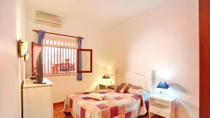 Een slaapkamer met een bed, nachtkastjes, een bureau, een stoel, een raam met tralies en een airconditioner.