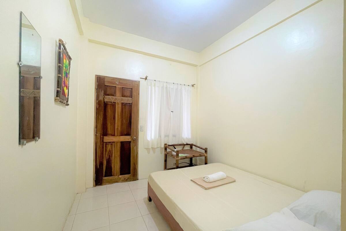 Une petite pièce propre, avec un lit, une porte en bois, une chaise et un miroir au mur.