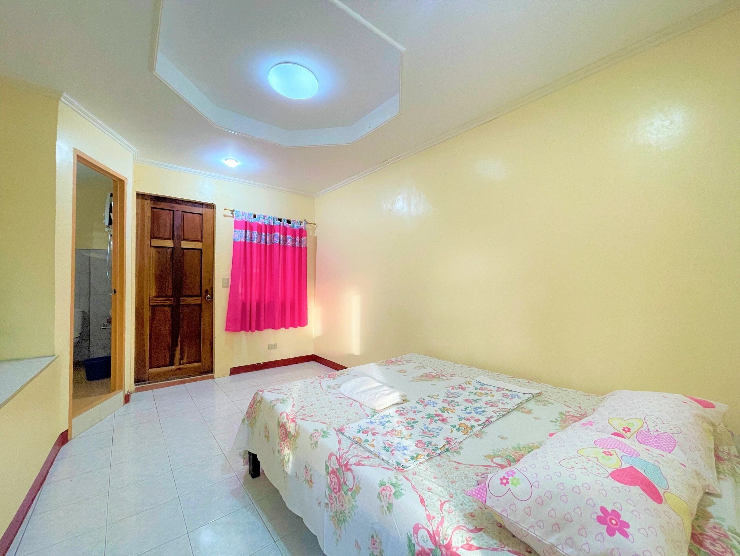 A bedroom with a bed, a door, a window with curtains, and a ceiling light.