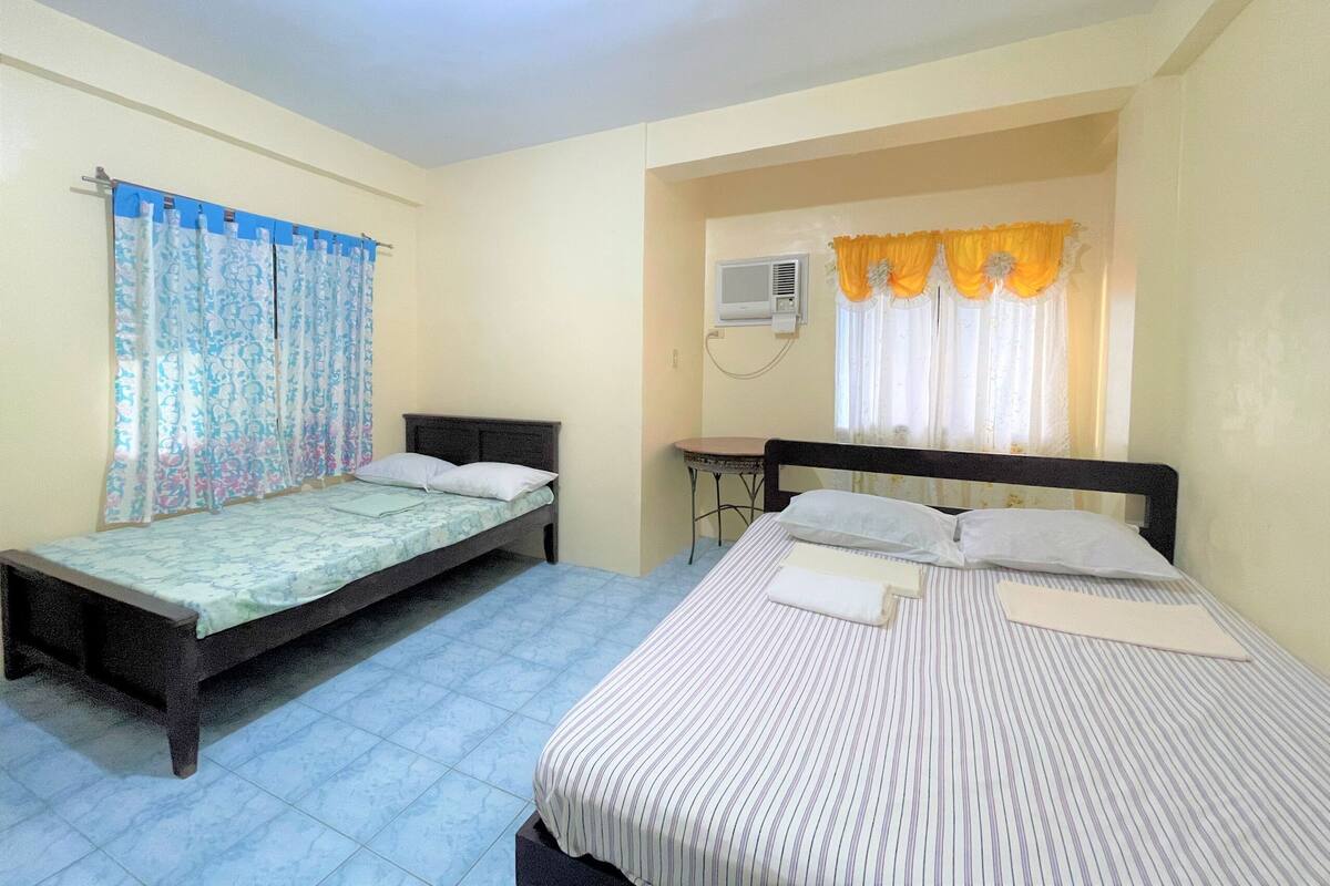 Une chambre avec deux lits simples, une petite table, un appareil de climatisation et une fenêtre avec des rideaux.