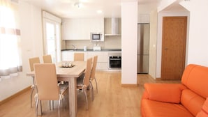A modern kitchen with white cabinets, a microwave, and a refrigerator. There is a dining table with chairs and an orange couch in the living area.
