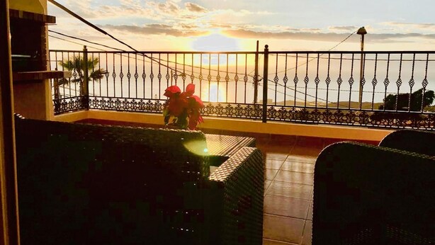 Un balcón con vistas a la puesta de sol sobre el mar, una barandilla y una pequeña mesa con flores.