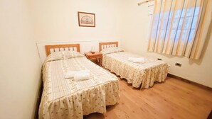 Una habitación con dos camas individuales, cabeceros de madera, una mesita de noche con lámpara y una ventana con persianas verticales.