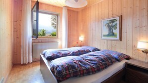 A wooden cabin room with two beds, a window with a view, a radiator, and a painting on the wall.
