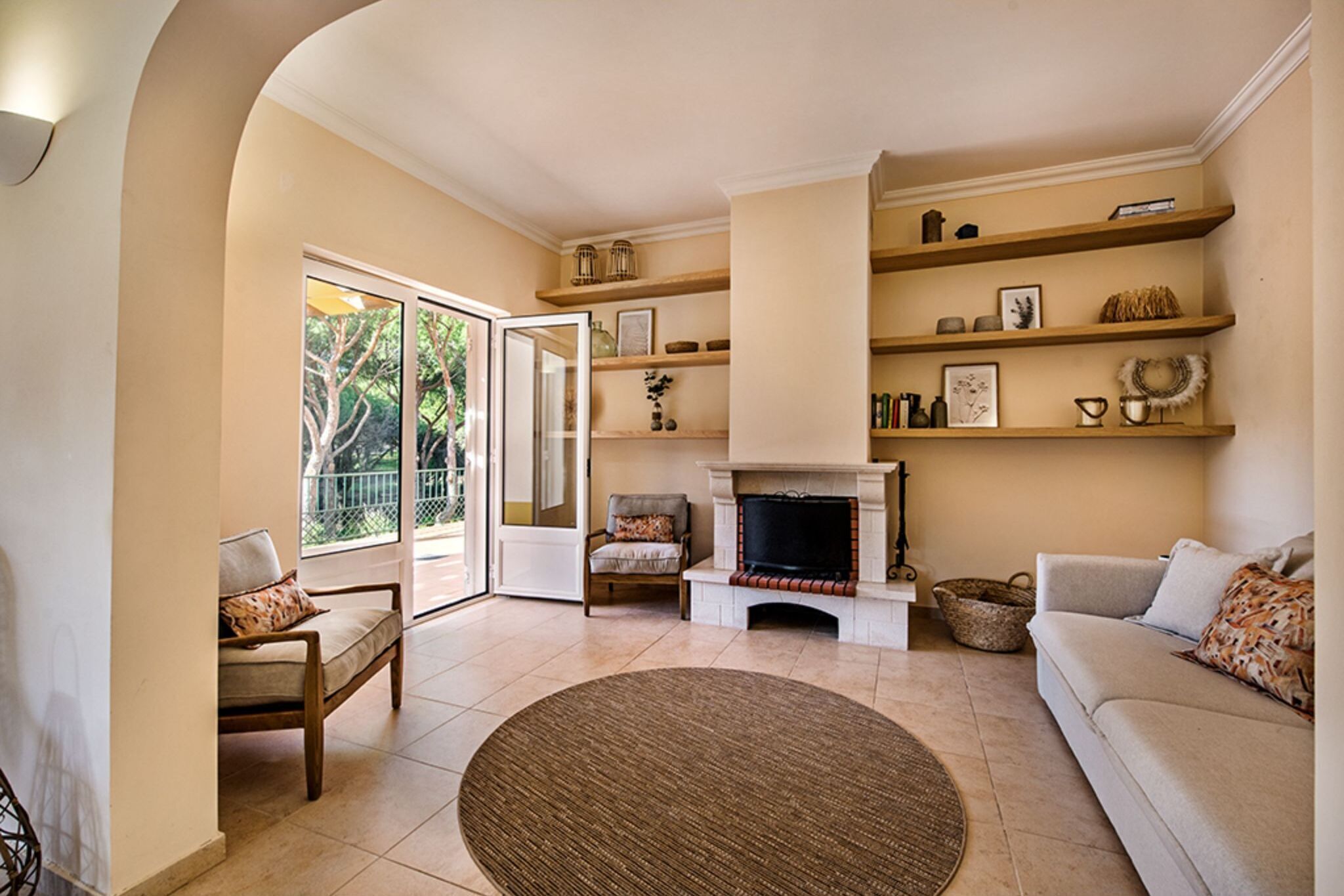 A living room with a fireplace, a sofa, a chair, and a television. There are shelves with decorative items and a view of the outdoors through a glass door.