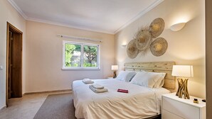 A neatly arranged bedroom with a large bed, bedside lamps, a wooden headboard, and a window with a view of greenery. - Large villa with swimming pool within walking distance to the beach (Castro Marim)