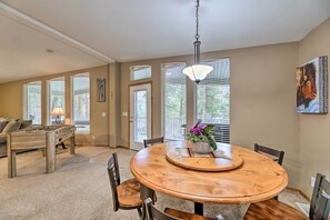 A dining area with a round wooden table, chairs, and a chandelier. There is a sofa with cushions in the background and a window with blinds.