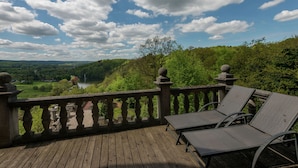 Una terraza de madera con barandilla de piedra que ofrece vistas panorámicas a un paisaje con un lago y colinas.