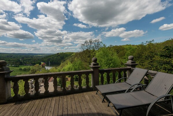 Une terrasse en bois, agrémentée d’une balustrade en pierre, offre une vue imprenable sur un paysage pittoresque, avec un lac et des collines.