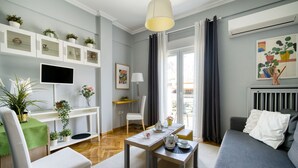 A modern living room with a sofa, a dining table, and a TV. There are potted plants on shelves and a window with curtains. - Snug apartment in Αθήνα with veranda (Αθήνα)