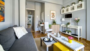 A modern living room with a grey sofa, a yellow bench, a coffee table, and a TV mounted on the wall. There are potted plants on shelves and a painting on the wall. - Snug apartment in Αθήνα with veranda (Αθήνα)