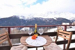 Een tafel met een fles en glazen op een balkon met uitzicht op besneeuwde bergen.