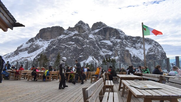 A wooden deck with tables and benches, overlooking a snow-covered mountain range.