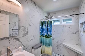 A bathroom with marble walls, a toilet, a sink, a shower curtain with a tropical design, and a window above the shower. - Breezy Monterey Apt: Walk to Downtown & Beach (Monterey)