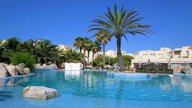 A clear blue pool with palm trees and rocks, surrounded by buildings.