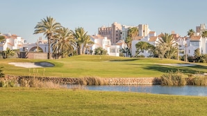 Un campo de golf con un estanque, palmeras y edificios blancos al fondo.