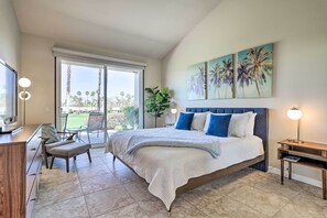 Un dormitorio con una cama grande, un escritorio, una silla y un televisor. Hay un ventanal corredera que da a un balcón con vistas a palmeras.