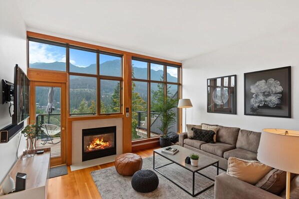 Ein modernes Wohnzimmer mit Kamin, großen Fenstern mit Blick auf die Berge, einer Couch und Kunstwerken an der Wand.