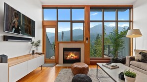 A modern living room with a fireplace, wooden flooring, a large window with a mountain view, a sofa, and a coffee table. - 3BR Townhouse • AC • Fireplace • Lake & Trail Views • 6-Min to Gondola (Whistler)