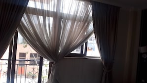 A room with a large window covered by sheer curtains, a metal balcony railing, and a view of buildings outside. - Mesfin Harar Hotel (Addis Ababa)