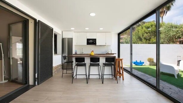 Eine moderne Küche mit weißen Schränken, einer schwarzen Arbeitsplatte und einer gelben Obstschale. Drei schwarze Stühle und ein Holzschemel sind ebenfalls vorhanden. Die Küche verfügt über eine große Glasschiebetür, die zu einem Garten mit einer weißen Badewanne und einem blauen Wasserspiel führt.