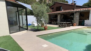 Ein moderner Poolbereich im Freien mit klarem Wasser, einem Glastür-Eingang und einer Terrasse mit Tisch und Stühlen.
