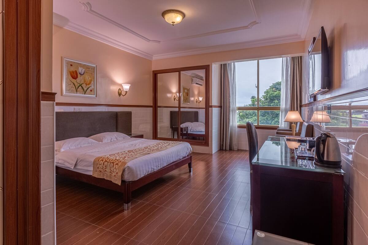 A hotel room with a bed, a desk with a kettle, a TV, and a large window with a view.