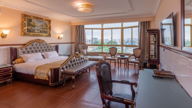A luxurious hotel room with a large bed, a sitting area, a desk, and a view of the city through large windows. - L'Etoile d'Or Hotel London (Libreville)