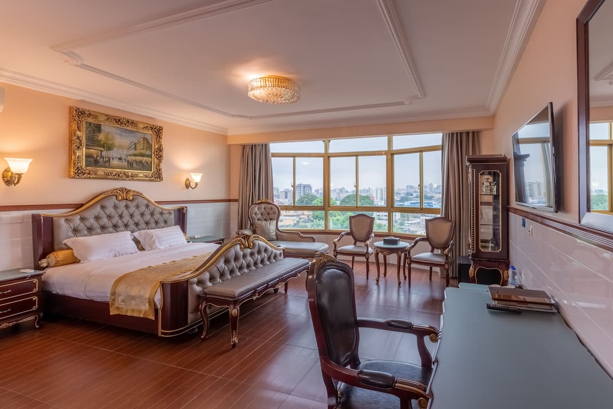 A luxurious hotel room with a large bed, a sitting area, a desk, and a view of the city through large windows.