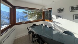 A modern dining room with a large glass table, black chairs, and a view of snow-covered mountains through large windows.