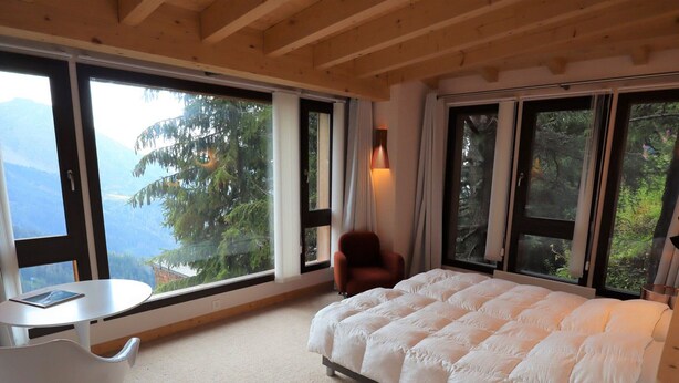 A bedroom with a bed, a chair, a table, and large windows offering a view of trees and mountains.