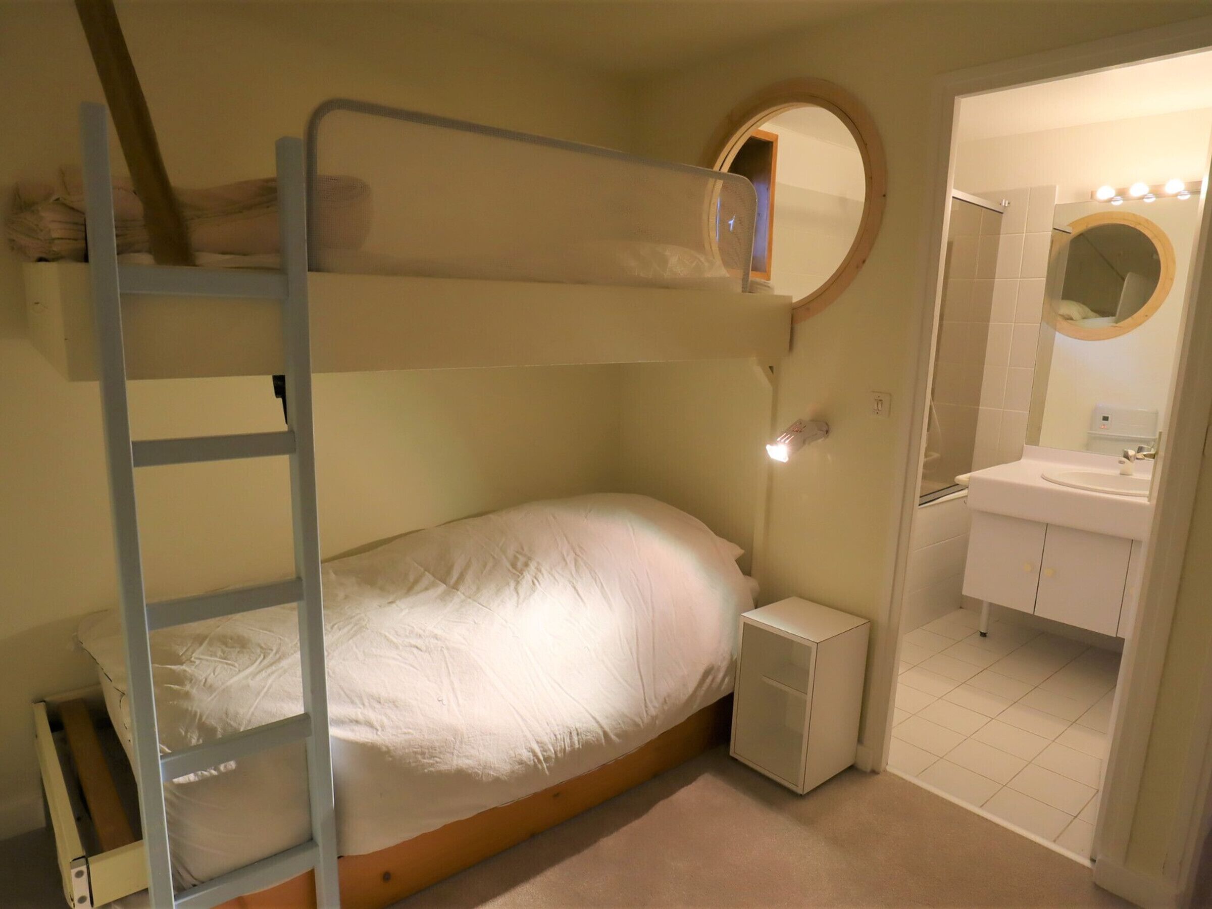 Uma cama beliche com escada, uma mesinha de cabeceira e um pequeno armário. Há um espelho redondo acima da pia e uma porta que dá para outro quarto.