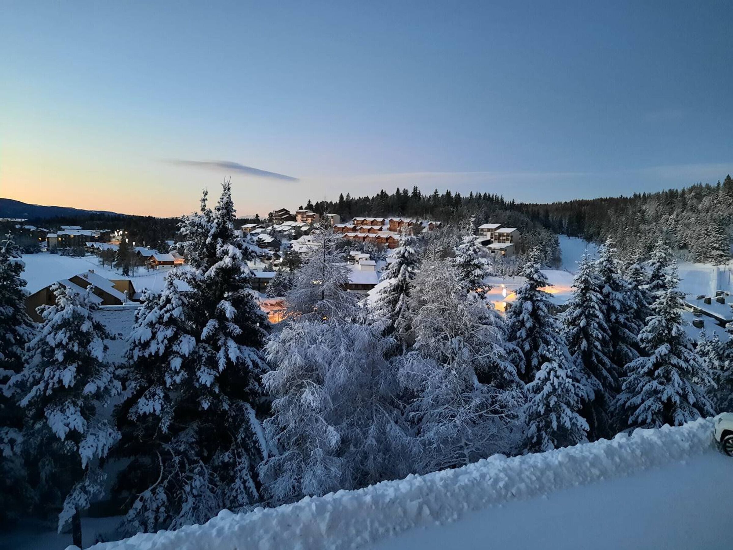 A snow-covered landscape with a cluster of buildings, likely a resort or lodge, surrounded by trees and a distant mountain range.