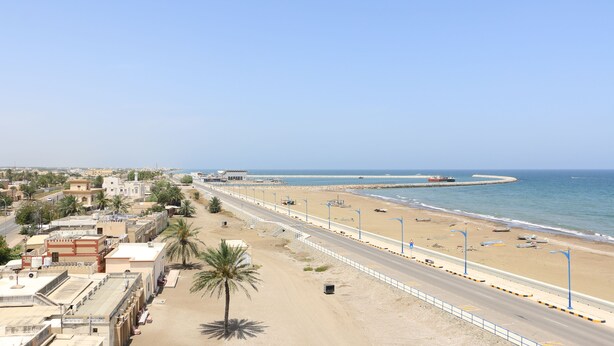 A coastal road with a beach on one side, lined with palm trees and buildings. - MARINA SOHAR HOTEL (Sohar)