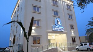 A multi-story hotel with a prominent sailboat logo on the facade, a staircase leading to the entrance, and a parking area with cars. - MARINA SOHAR HOTEL (Sohar)