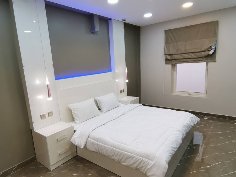 A modern bedroom with a large bed, bedside tables, a wall-mounted light, and a window with a blind.