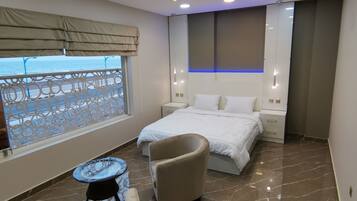 Una habitación de hotel moderna con una cama grande, un sillón curvo, una pequeña mesa redonda y vista al mar a través de una ventana con diseño.